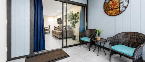 A living area with a sliding glass door leading to a balcony, wicker furniture, a small table, and a decorative wall piece.