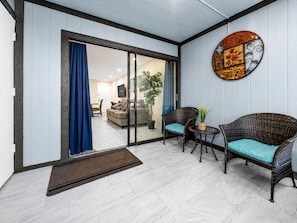Espaço de estar com porta de vidro deslizante para uma varanda, móveis de vime, uma mesa pequena e um objeto decorativo na parede.