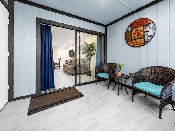 A living area with a sliding glass door leading to a balcony, wicker furniture, a small table, and a decorative wall piece.