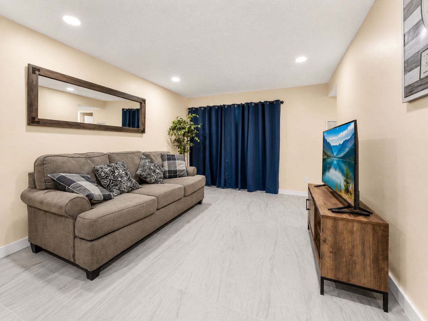 Una sala de estar con un sofá, un televisor sobre un mueble de madera, un espejo en la pared y una planta.
