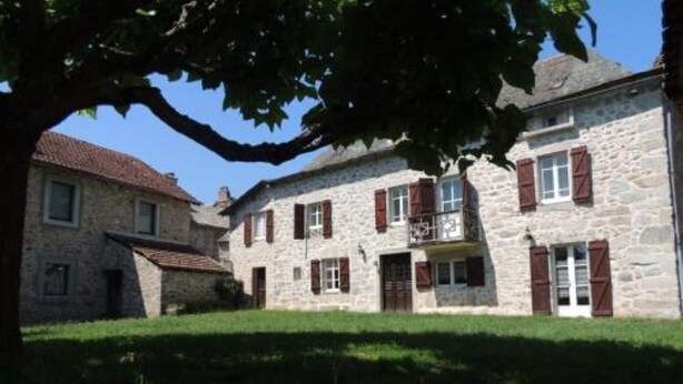 A stone building with red shutters, a balcony, and a grassy courtyard. - L'oustal Bergounhou (Le Bas-Ségala)