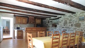 A dining area with a long table and chairs, a kitchen with stone walls, wooden cabinets, and a stone fireplace. - L'oustal Bergounhou (Le Bas-Ségala)