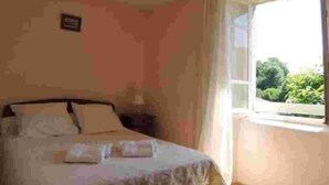 A bedroom with a bed, a nightstand, and a window with a view of greenery outside. - L'oustal Bergounhou (Le Bas-Ségala)
