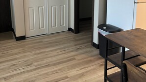 Une pièce avec un plancher en bois, une porte blanche, une poubelle noire et une table en bois avec une chaise.