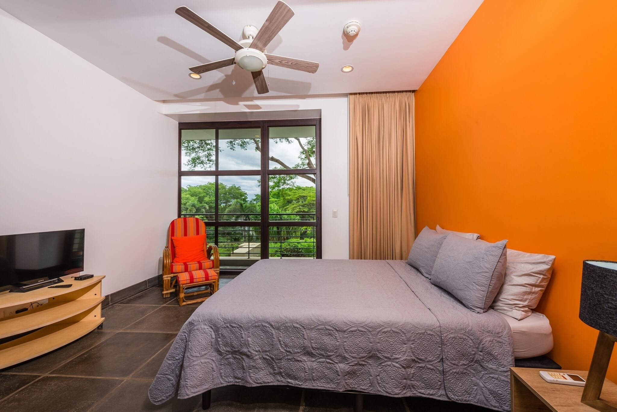 A bedroom with a bed, a TV, a ceiling fan, and a window with a view of greenery.