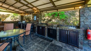 Un patio exterior con una barbacoa de piedra y armarios empotrados, rodeado por una zona cubierta con techo metálico.