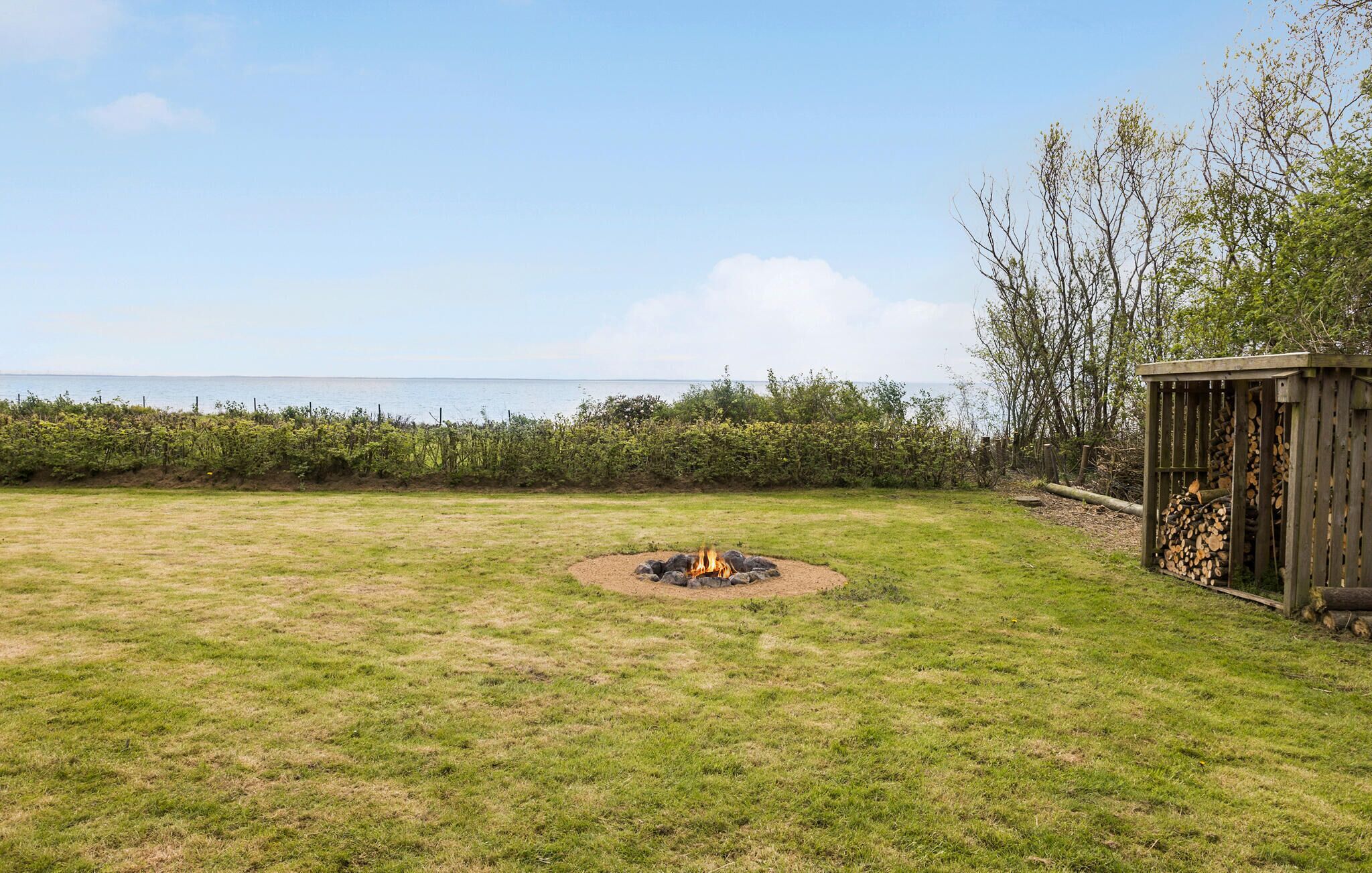 A grassy outdoor area with a fire pit in the center, a wooden structure on the right, and a view of the sea in the background.
