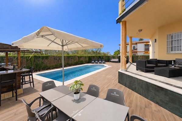Un espace aménagé au bord de la piscine, comprenant une table, des chaises, un grand parasol et des sièges d’extérieur.