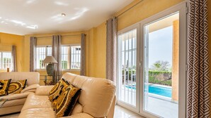 A spacious living room with a beige leather sofa, patterned cushions, and a view of a swimming pool through large sliding glass doors.