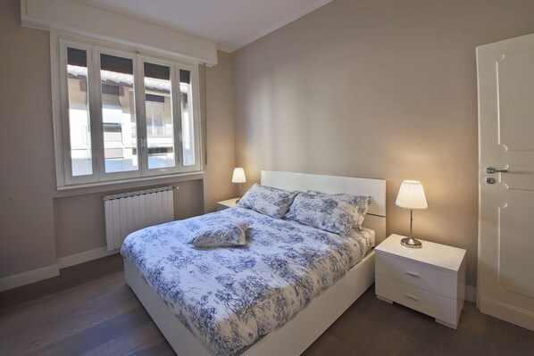 A bedroom with a bed, two bedside tables, a window, and a radiator.