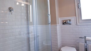Une salle de bain avec des murs recouverts de carreaux blancs, une douche en verre, des toilettes et un porte-serviettes.