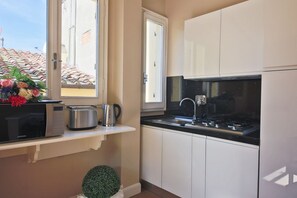 Une cuisine compacte équipée d’un four à micro-ondes, d’une bouilloire et d’une cuisinière, une fenêtre donnant sur un toit recouvert de tuiles et une petite plante en pot.