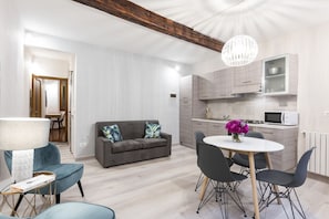 Un salon moderne comprenant un canapé, une table à manger et des chaises, une cuisine avec des placards et un four à micro-ondes, et une poutre en bois au plafond.
