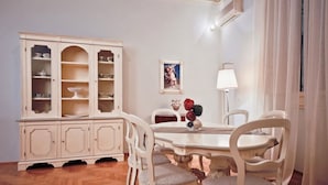 Un comedor con una mesa blanca, sillas, un aparador con vajilla, una lámpara y un cuadro en la pared.