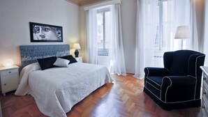 Un dormitorio con cama, una silla, una mesita de noche, una lámpara y un ventanal con cortinas.