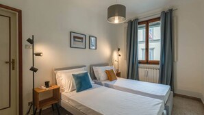 Een slaapkamer met twee bedden, nachtlampjes, een vloerlamp en een raam met gordijnen.