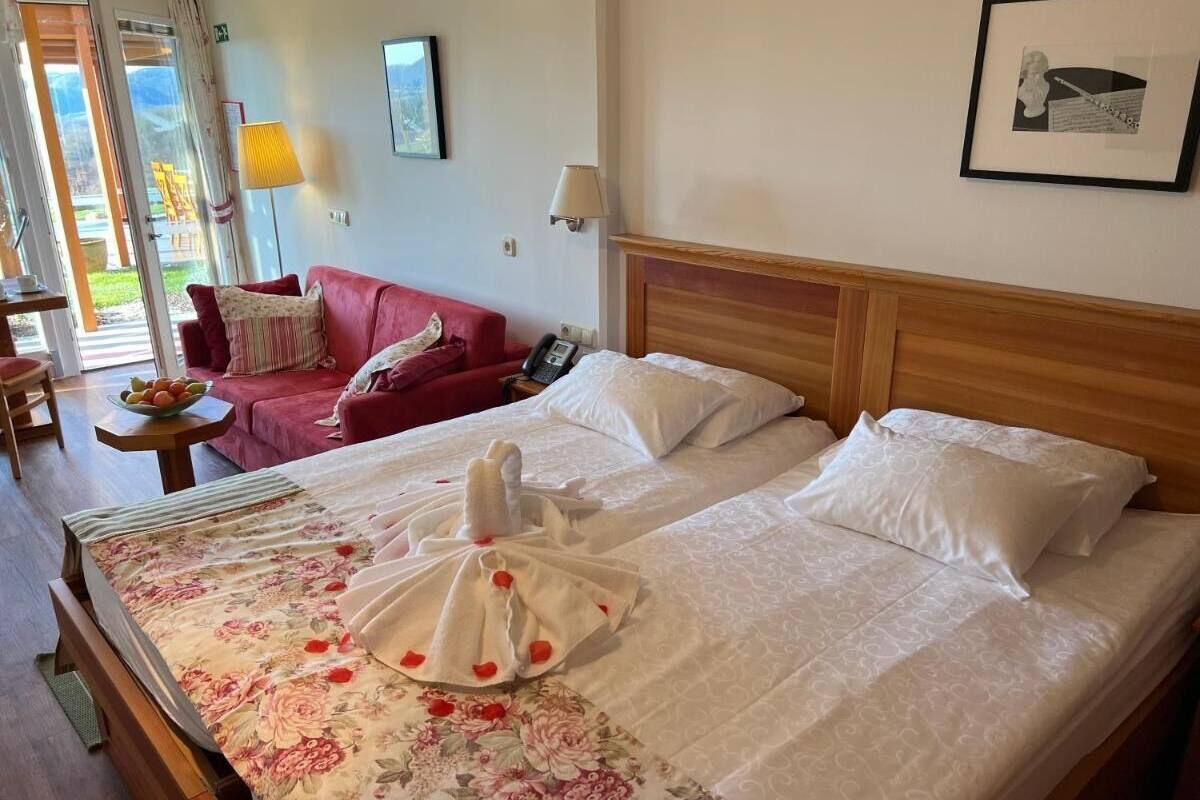 Une chambre d’hôtel avec un grand lit, un canapé rouge, une petite table sur laquelle se trouve un panier de fruits, et une vue sur l’extérieur grâce à une porte vitrée.