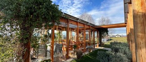 Un área de estar al aire libre con techo, mesas y sillas, rodeada de vegetación y una estructura de madera.