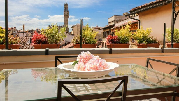 A glass table with a white plate of flowers, a chair, and a view of buildings with a tower in the background.