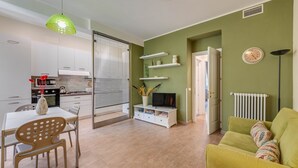 Una cocina moderna con armarios blancos, una mesa de comedor con sillas, un televisor sobre un mueble blanco, y un sofá verde en una sala de estar con un radiador y un reloj en la pared.