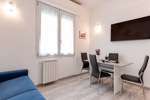 A modern room with a blue sofa, a dining table with four chairs, a TV mounted on the wall, and a window with curtains. - Frullhouse - Sweet Dreams in Florence (Coverciano)