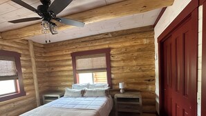 Un dormitorio de cabaña de madera con cama, mesitas de noche, ventilador de techo, ventana con persianas y una puerta roja.