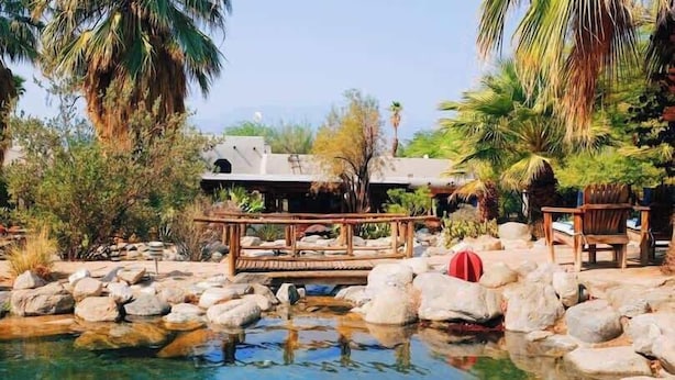 A tranquil outdoor pool area with rocks, a wooden deck, and palm trees. - Lockout The Oasis | Natural Mineral Hot Springs on 107 Acres (Desert Hot Springs)