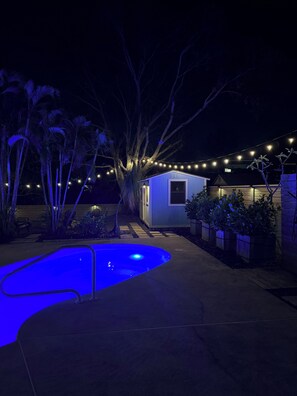 Ein Schwimmbadbereich im Freien bei Nacht mit Lichterketten, einem kleinen Schuppen und einem Holzzaun.