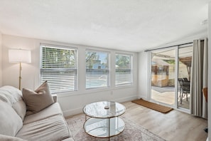 Ein modernes Wohnzimmer mit einer Couch, einem Glastisch und großen Fenstern mit Jalousien.