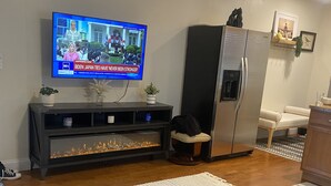 Ett vardagsrum med en tv-apparat som visar ett nyhetsprogram, ett träbord med en skål med frukt, en soffa i mörk färg, ett kylskåp och en liten växt på hyllan.