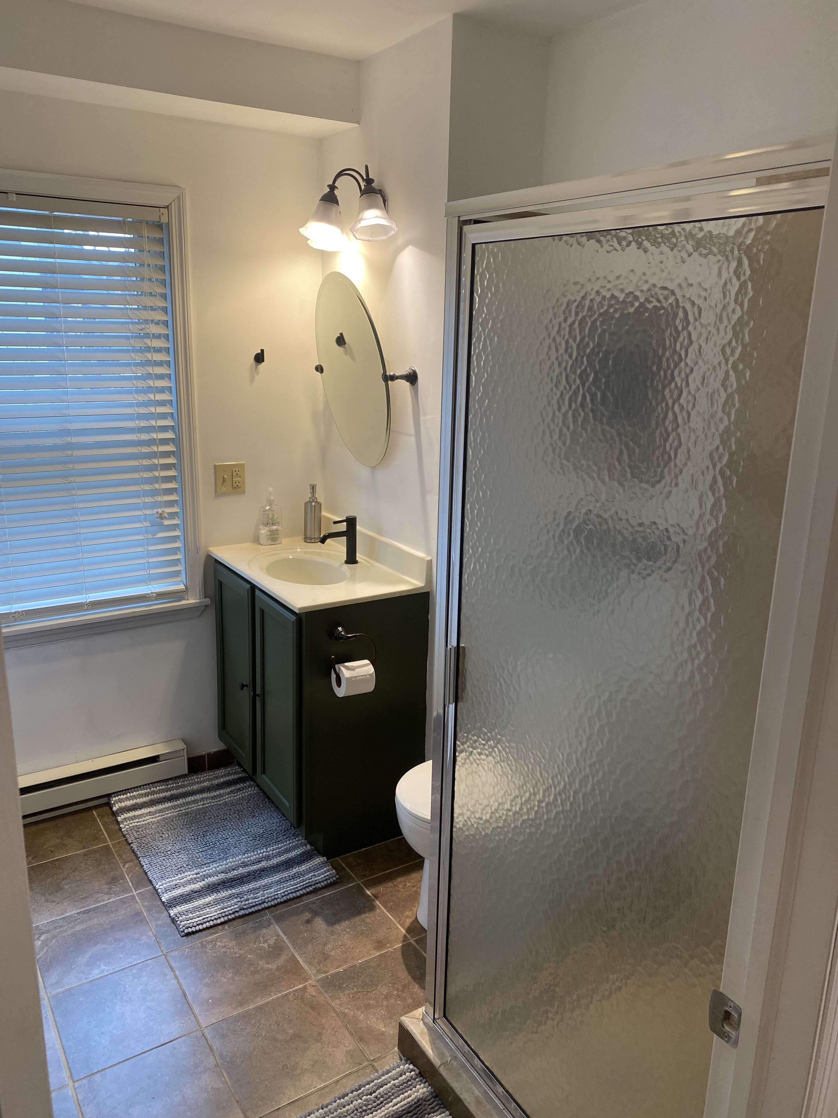 Une salle de bain avec des toilettes, un lavabo et un miroir. Il y a une fenêtre avec des stores, un porte-serviettes et une douche avec une porte en verre.