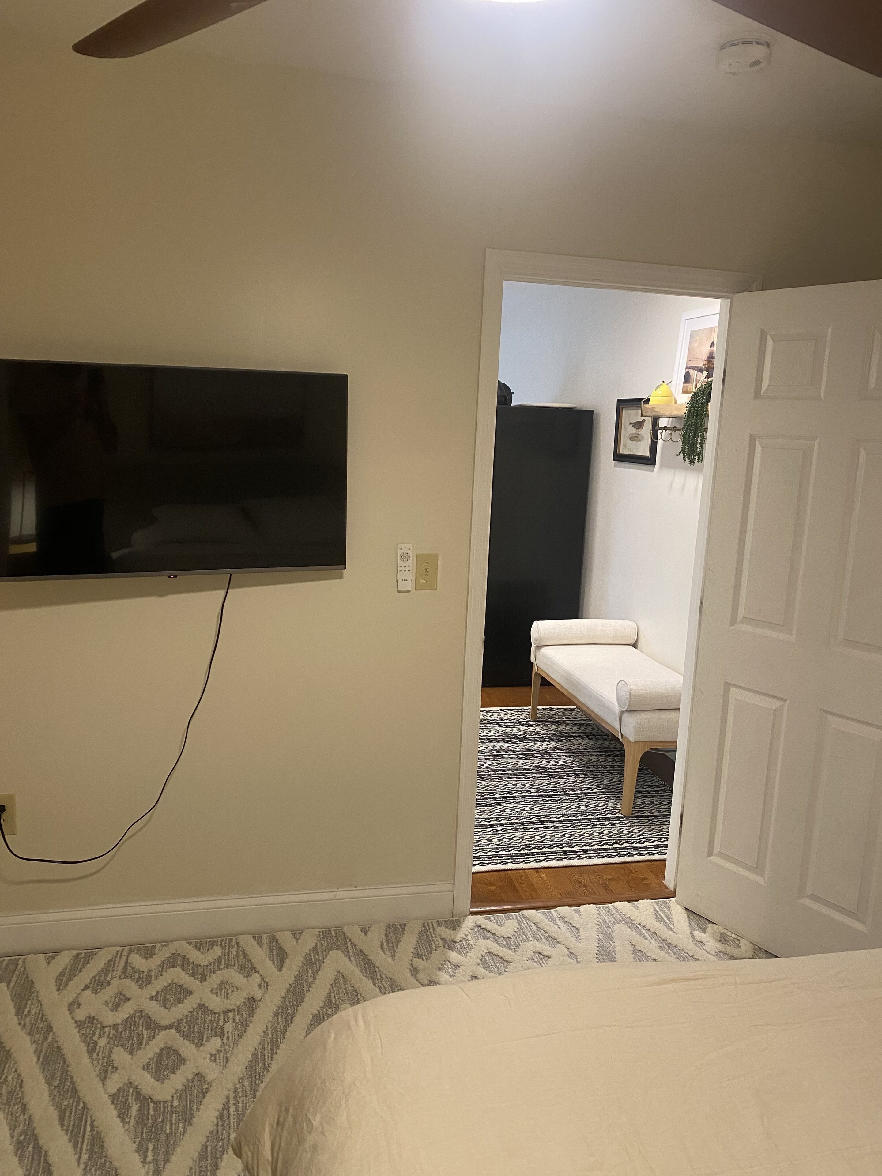 Un dormitorio con una cama, un televisor montado en la pared, una puerta que da a otra habitación y una alfombra estampada.