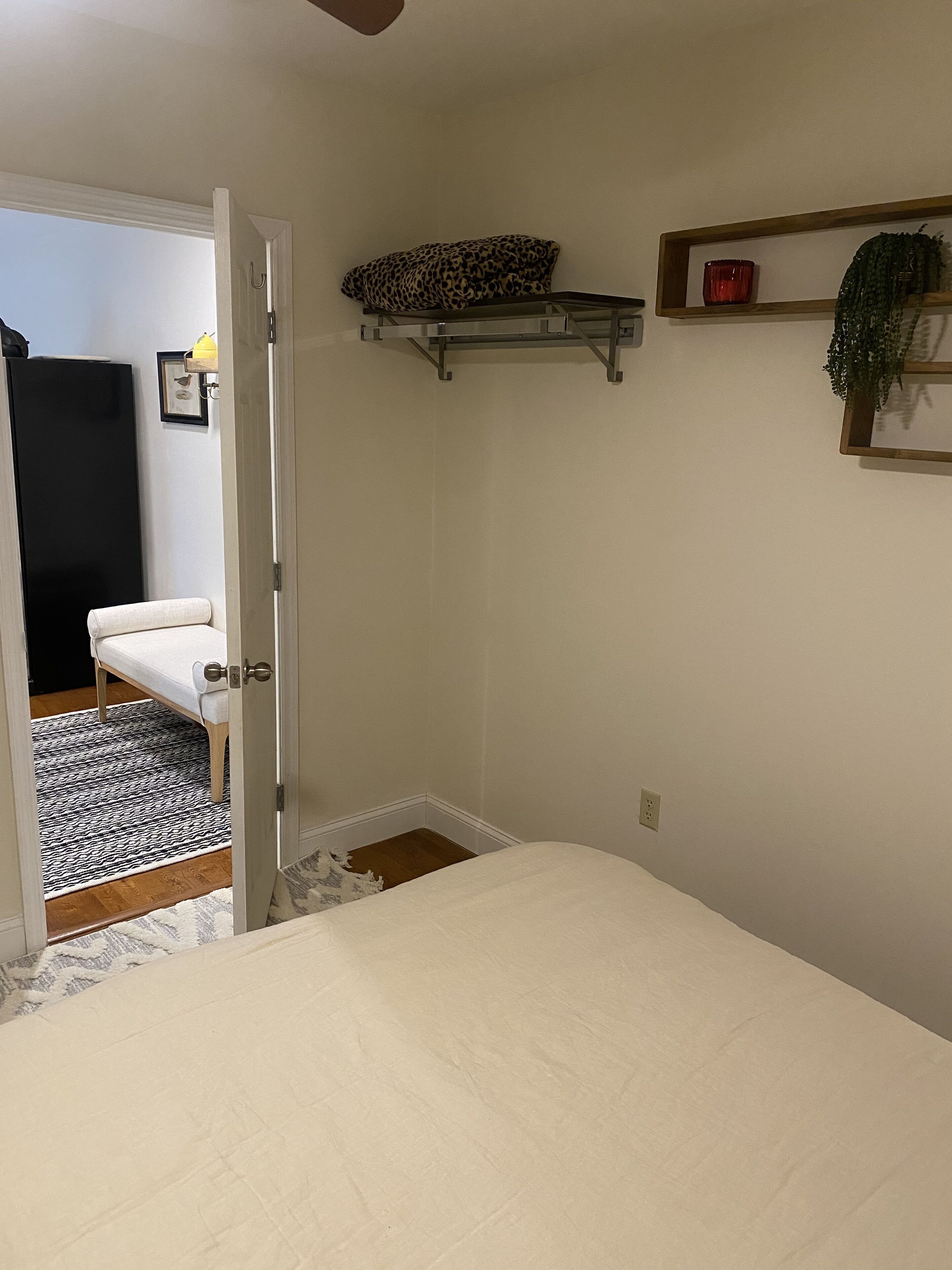 Un dormitorio con una cama, una puerta que da a otra habitación, un estante pequeño con una planta y un cuadro enmarcado en la pared.