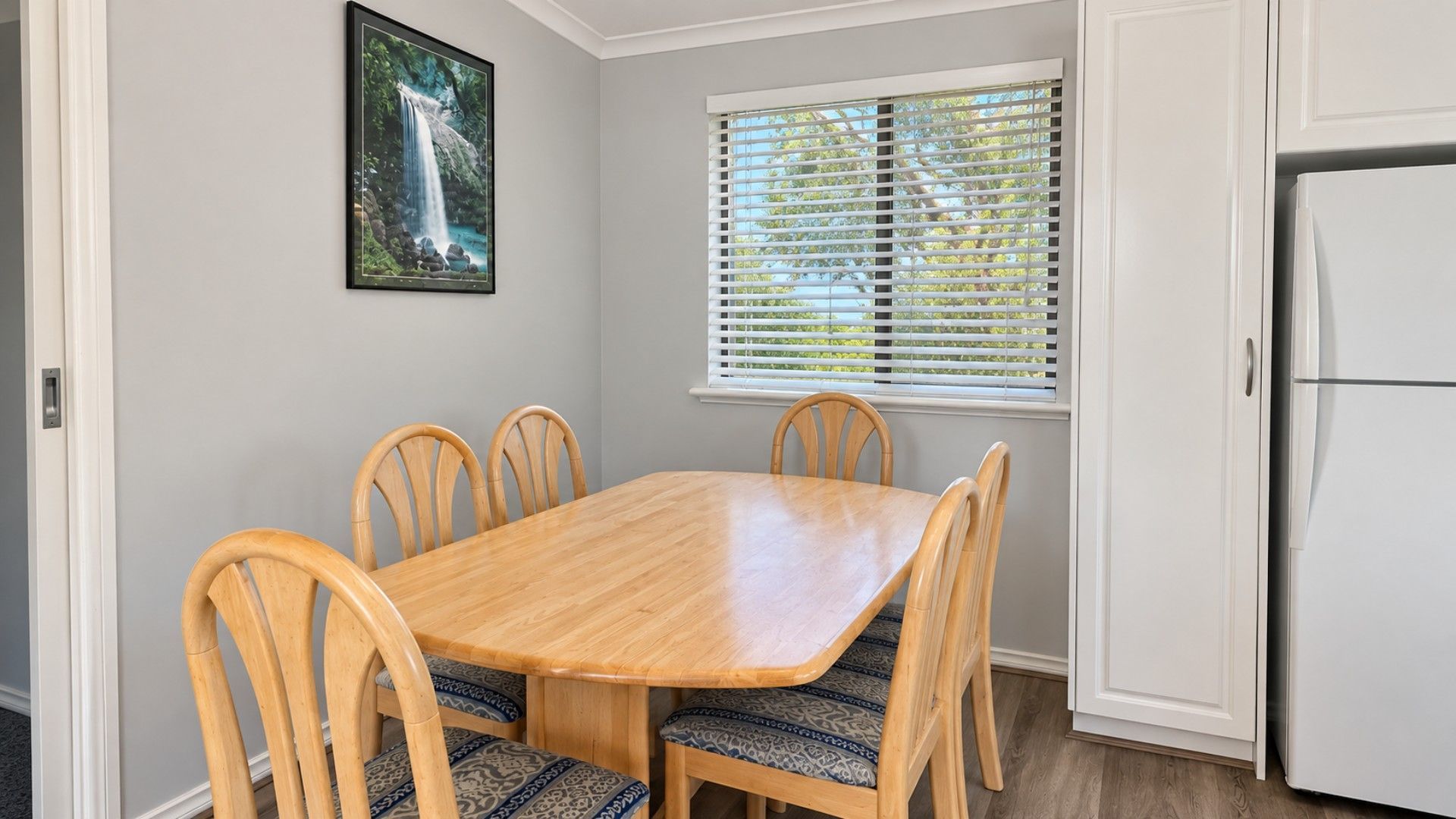 A dining area with a wooden table and four chairs, a window with blinds, and a refrigerator.