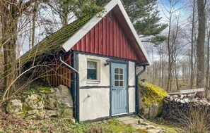 Une petite maison en bois d’un seul étage, avec un toit rouge et des murs blancs, entourée d’arbres et d’un mur de pierres.