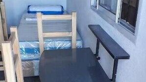 Una habitación pequeña con una mesa de madera y dos sillas, una cama litera con colchón azul y una ventana con persianas.