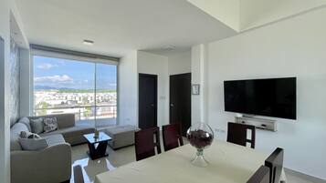 Una sala de estar moderna con comedor, ventanas amplias, televisor de pantalla plana y vistas a un paisaje urbano.