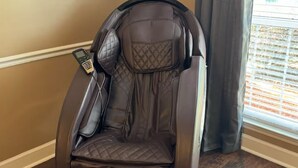 A massage chair with a control panel, positioned in a room with wooden flooring and a window with blinds.