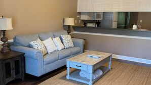 A living room with a grey sofa, two side tables, a coffee table, a ceiling fan, and a kitchen area in the background.