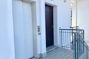 A white building with a dark door, a metal railing, and a tiled floor.