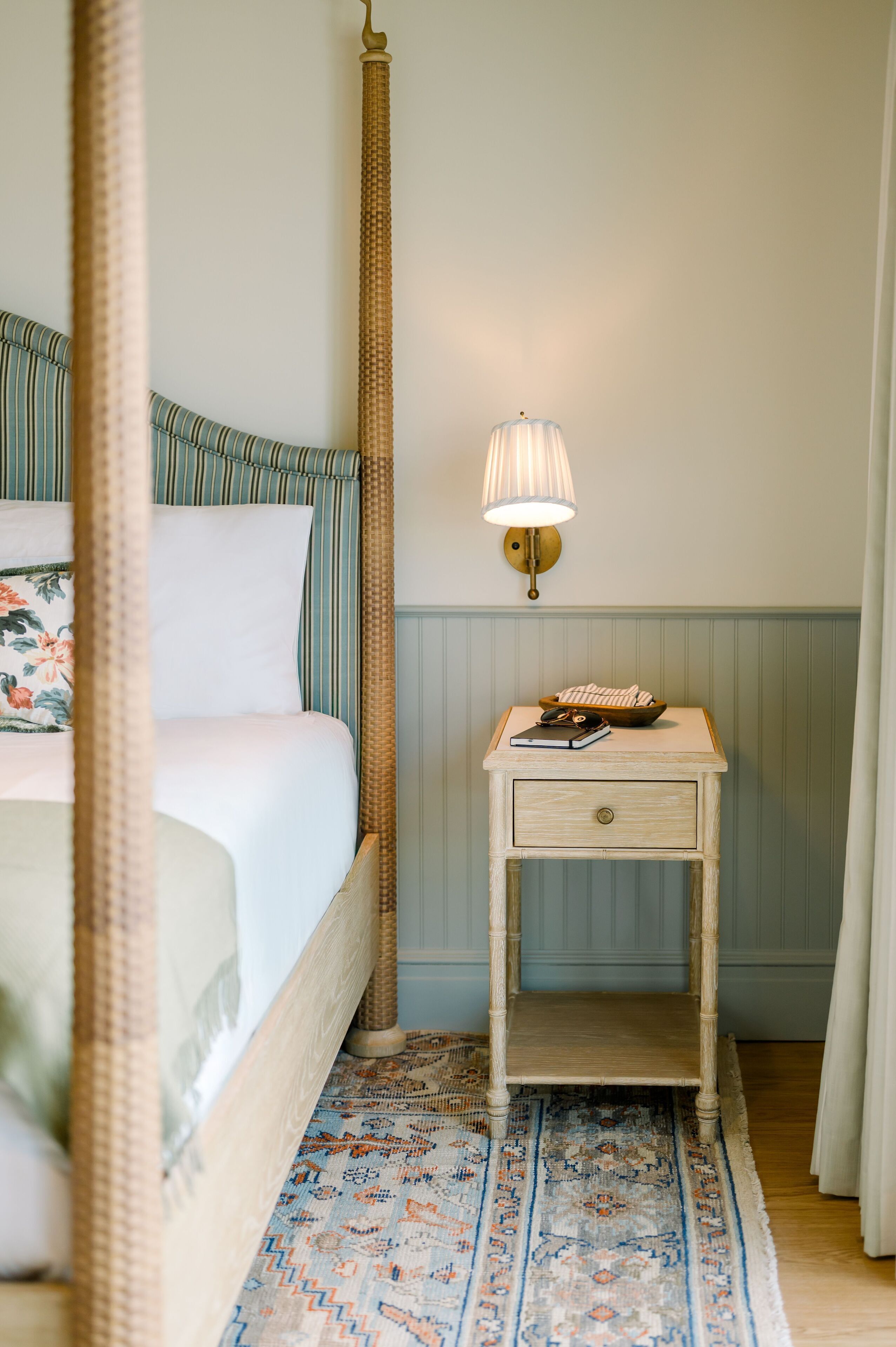A four-poster bed with a floral pillow, a bedside table with a lamp, and a patterned rug.