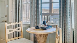 A room with a round table, wicker chair, and a view of the beach through a window with patterned curtains. - Oceanside King Suite | Near Pool + Private Beach (Duck)