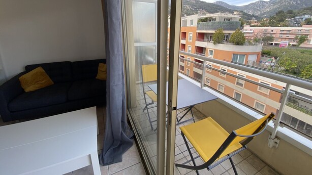 Un balcon avec un canapé noir et des coussins jaunes, une porte vitrée et une vue sur des immeubles et des montagnes.
