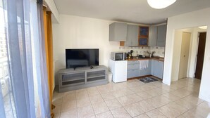 Un appartement moderne doté d’un espace de vie comprenant un téléviseur à écran plat installé sur un meuble de rangement gris, une cuisine compacte avec des placards gris et un four à micro-ondes, ainsi qu’un sol carrelé.