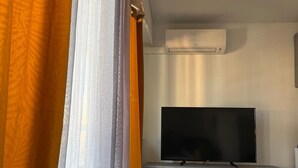 Un salon moderne avec un téléviseur à écran plat posé sur un meuble de rangement gris, un climatiseur blanc fixé au mur et une grande fenêtre ornée de rideaux orange.