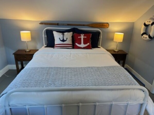 A neatly made bed with a white comforter and two pillows, one with an anchor design. There are two bedside tables with lamps, and a life preserver hangs on the wall.