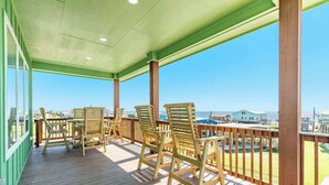 A covered outdoor deck with wooden chairs and a table, overlooking a coastal area with buildings and a clear sky. - New 5BR Beach House with Dual Decks and Fire Pit (Surfside Beach)