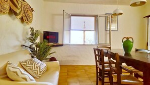 Ein Wohnzimmer mit Sofa, Fernseher, Esstisch mit Stühlen, einer Pflanze und einem Fenster mit Jalousien.