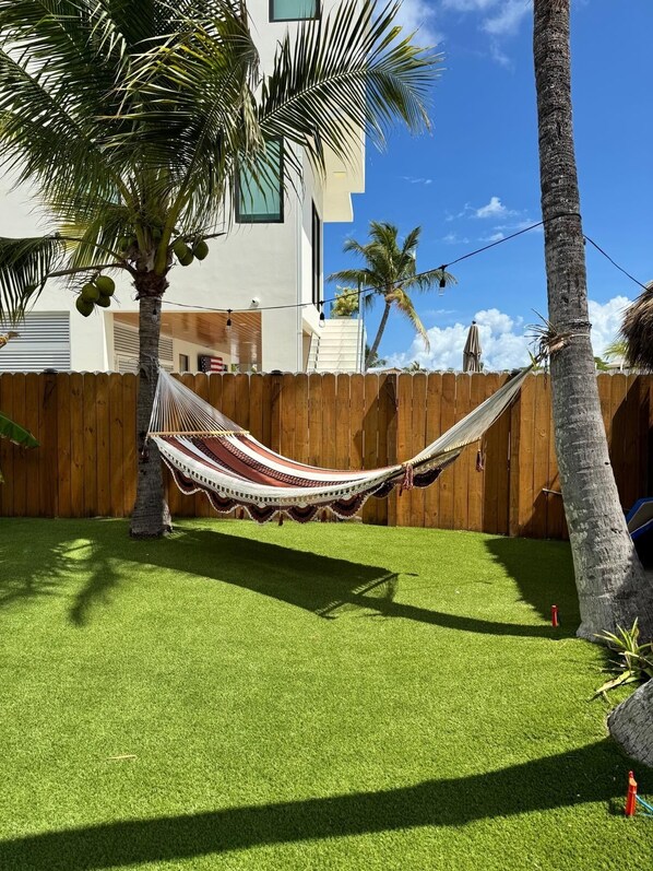 A hammock tied between two trees in a backyard with artificial grass, a wooden fence, and a building in the background.