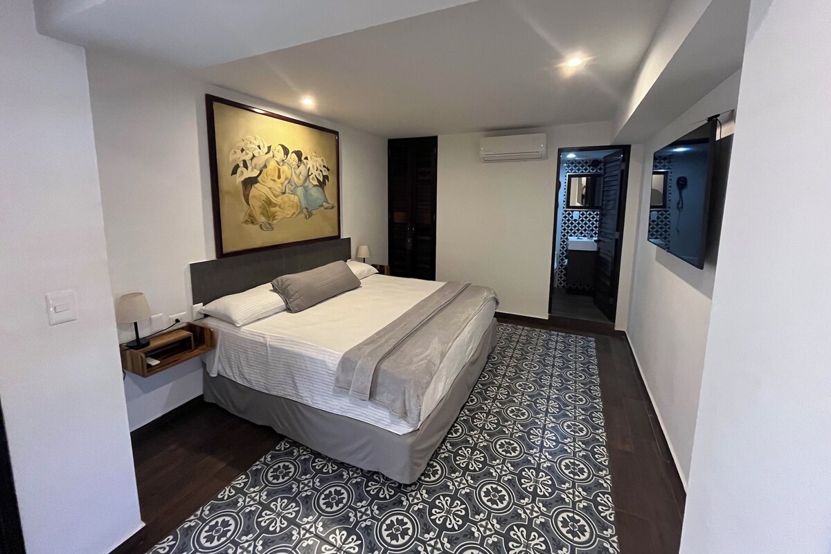 Una habitación de hotel moderna con una cama grande, un cuadro sobre la cama, una mesita de noche pequeña con una lámpara y un suelo estampado.